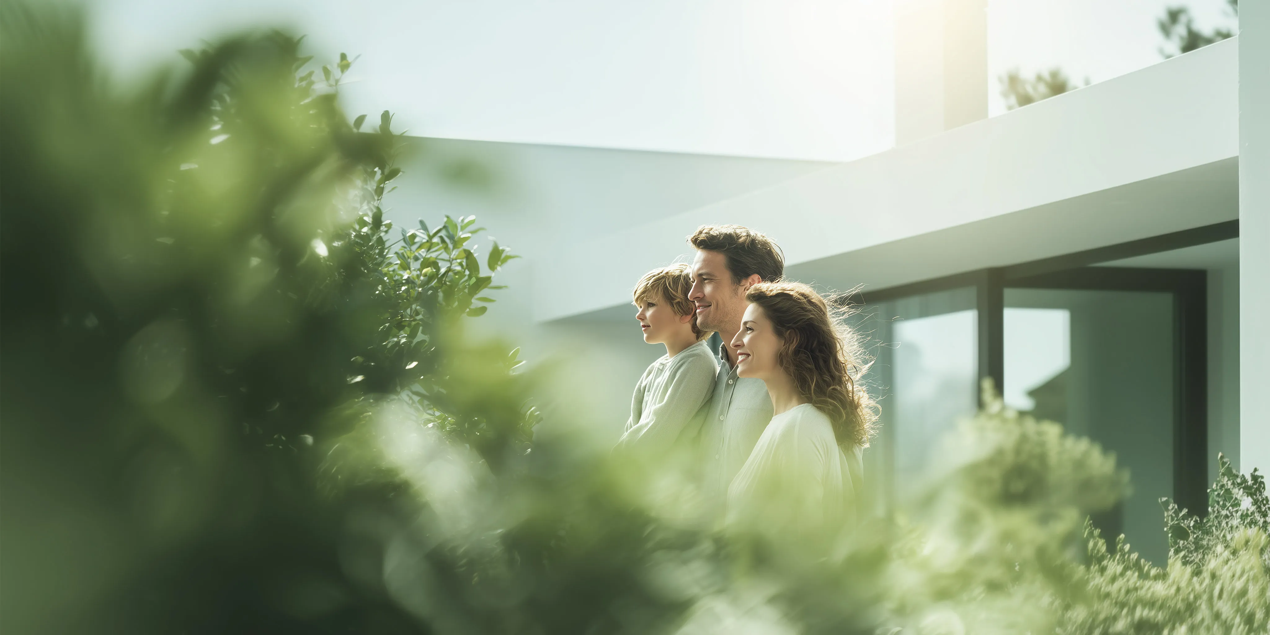 Smiling family of three standing outside modern house, surrounded by greenery on a sunny day.
