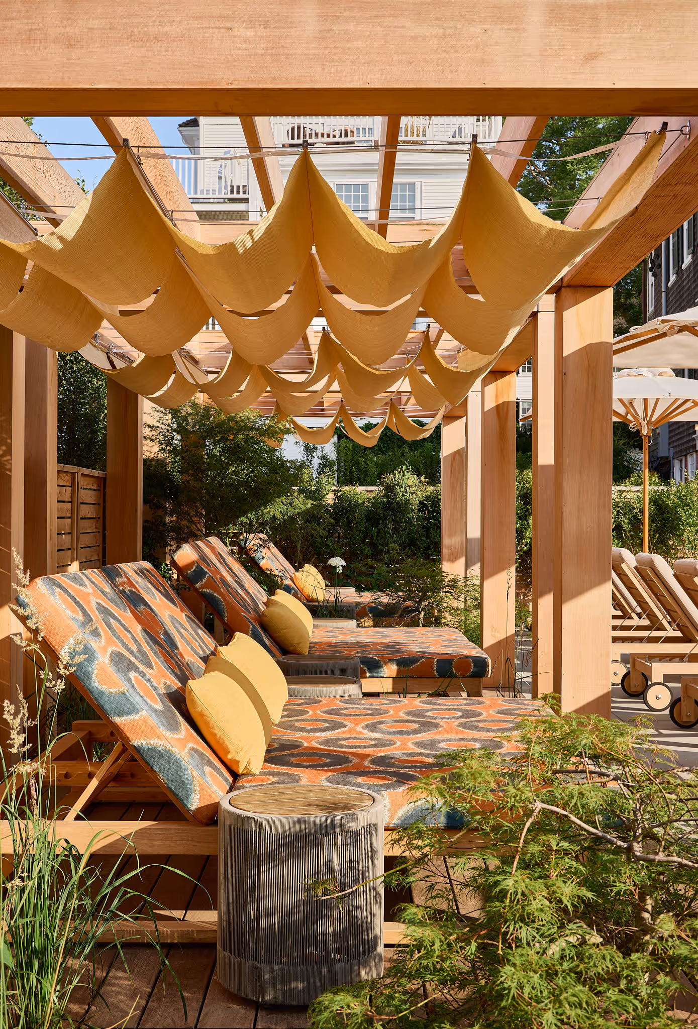 Outdoor lounge area with patterned reclining chairs, yellow cushions, side tables, and fabric canopy under a wooden pergola.