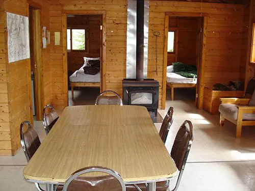 Interior of the Confusion Lake outpost cabin showing a dining table, chairs, wood stove, and two bedroom doorways.