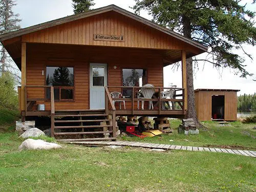Wilderness Air Escapes outpost cabin at Confusion Lake, with front veranda, patio chairs, wooden steps, and a small outbuilding amid spruce trees.
