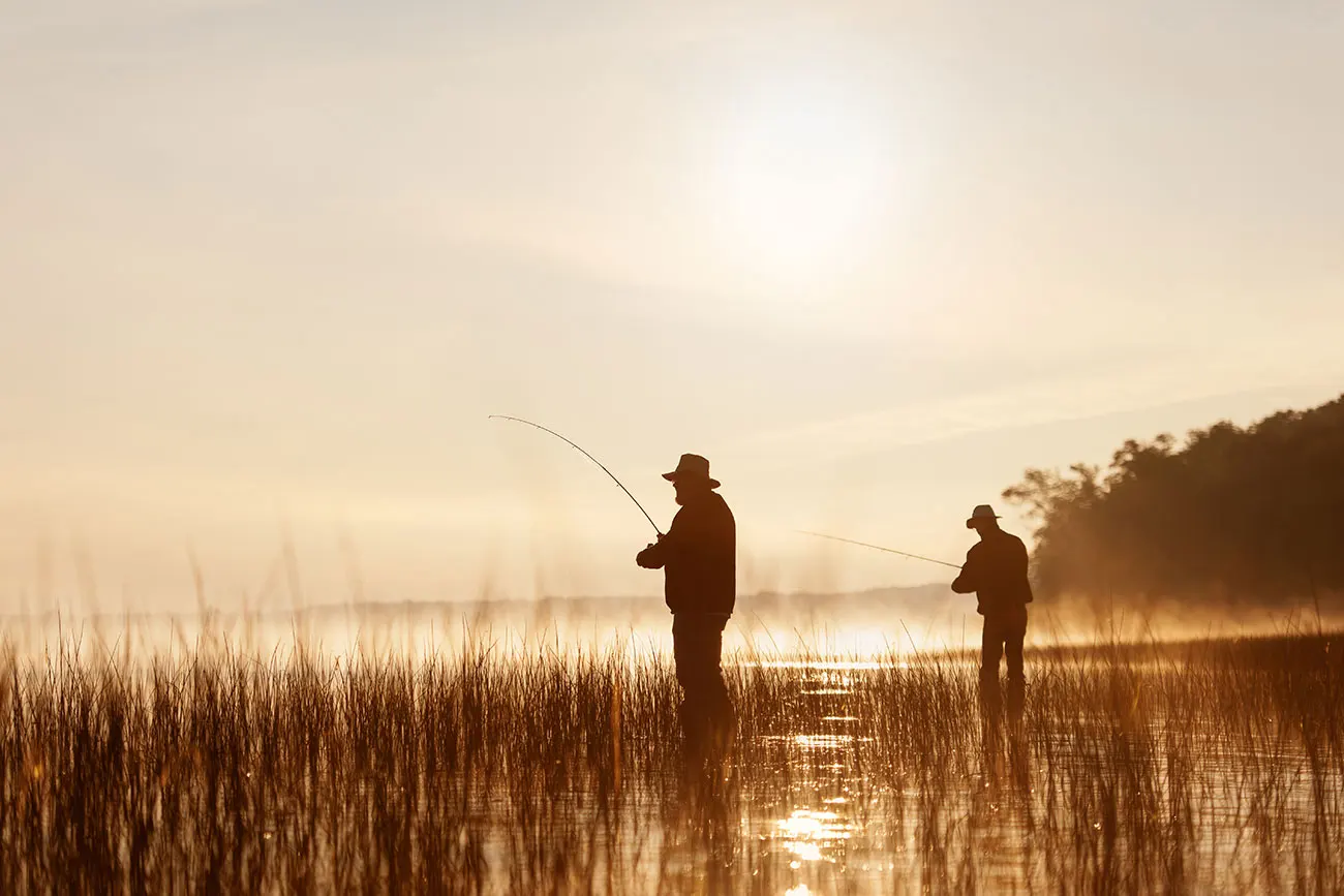 Two anglers casting fishing rods in a misty marsh at golden sunrise, silhouetted against glowing water and reed grass on Confusion Lake.