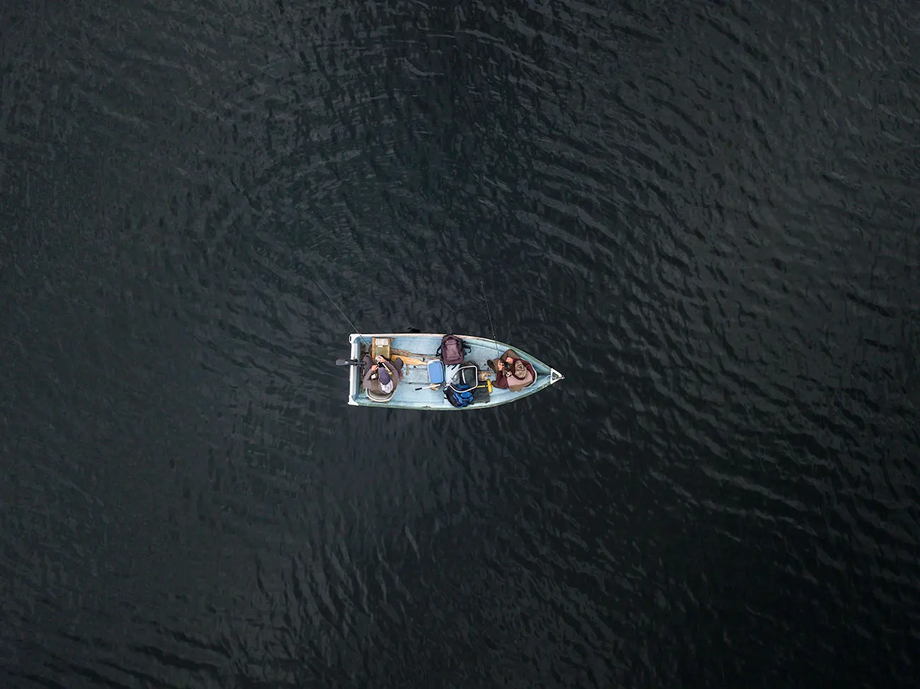 Aerial drone view of two anglers in a small aluminium fishing boat on dark open water, fishing rods extended.
