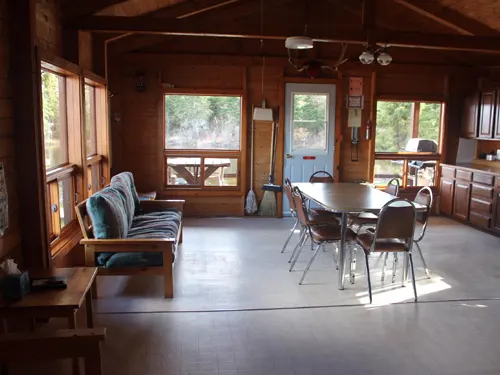Spacious log-walled cabin interior with dining table, folding chairs, a futon sofa, and large windows overlooking the forest.