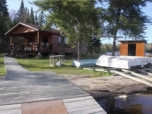Wooden dock leading to a Confusion Lake outpost cabin, with aluminum boats stored on shore and a small outbuilding nearby.
