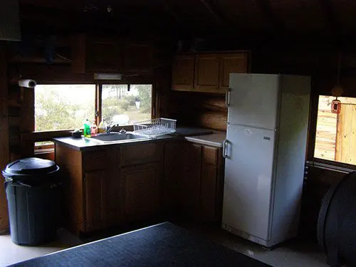 Interior of the Crooked Lake cook cabin kitchen, showing wood cabinets, white refrigerator, sink, and trash can.