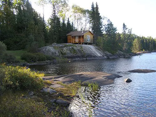 Yellow outpost cabin perched on a granite outcrop above Crooked Lake, framed by boreal forest of spruce and birch.