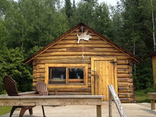 Restored 1954 trapper's cook cabin with log walls, moose antlers above the gable, and Adirondack chairs on a wooden deck.