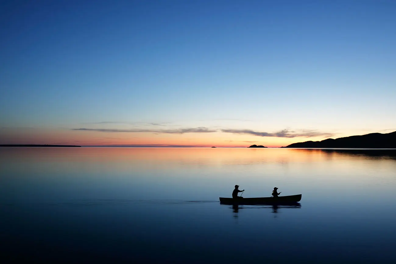 Two silhouetted figures paddle a canoe across glassy Crooked Lake at dusk, warm sunset hues reflected in the still water.