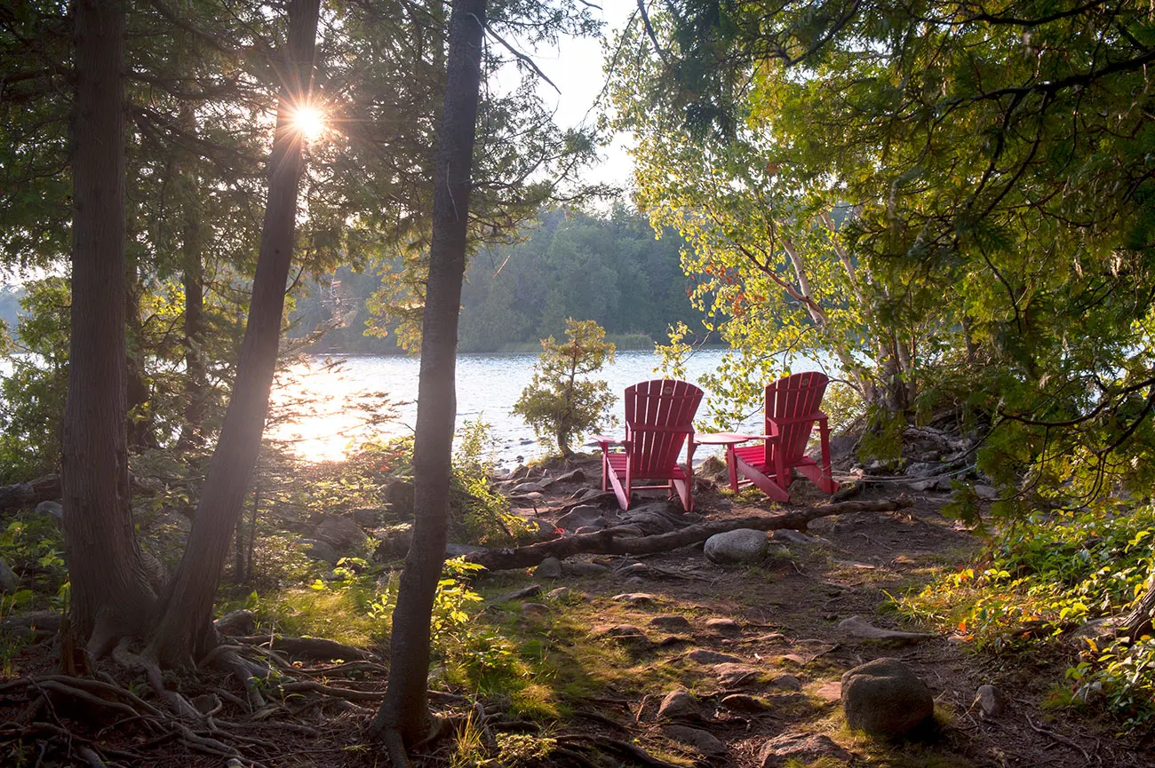 Two red Adirondack chairs on a rocky shoreline at Crooked Lake, framed by birch and pine as morning sun bursts through the trees.