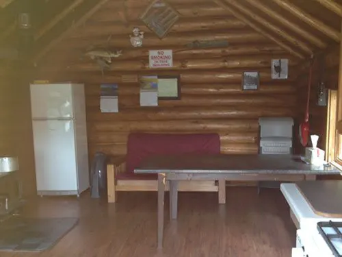 Interior of the Crooked Lake cook cabin, with log walls, a white fridge, red futon sofa, wood table, and a no-smoking sign.