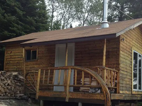 Log-sided outpost cabin with rustic branch railing deck, stacked firewood, brown shingle roof, and metal stovepipe, surrounded by forest.