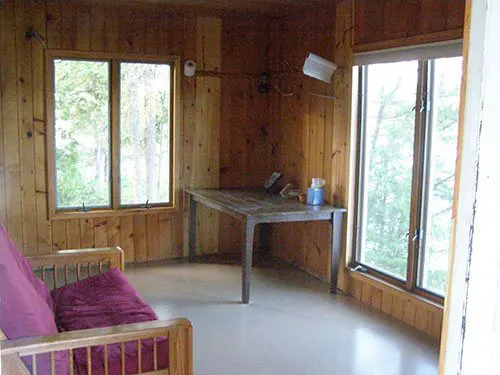 Interior of the Crooked Lake cook cabin showing knotty pine walls, a rustic wooden table, and a futon with purple cushions beside large windows.