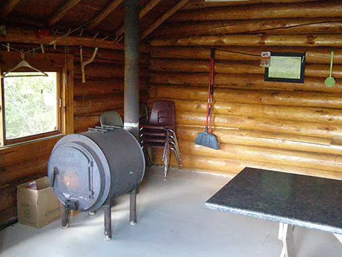 Interior of the Crooked Lake cook cabin showing a barrel wood stove, log walls, stacked chairs, broom, and a table.