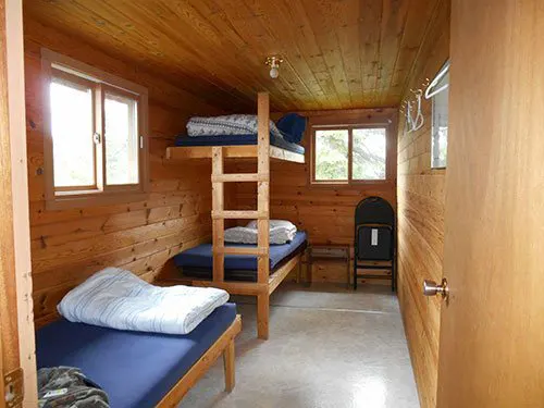 Interior of the Pilot Lake outpost cabin showing a single bed, bunk beds with ladder, pine-panelled walls, and two windows.