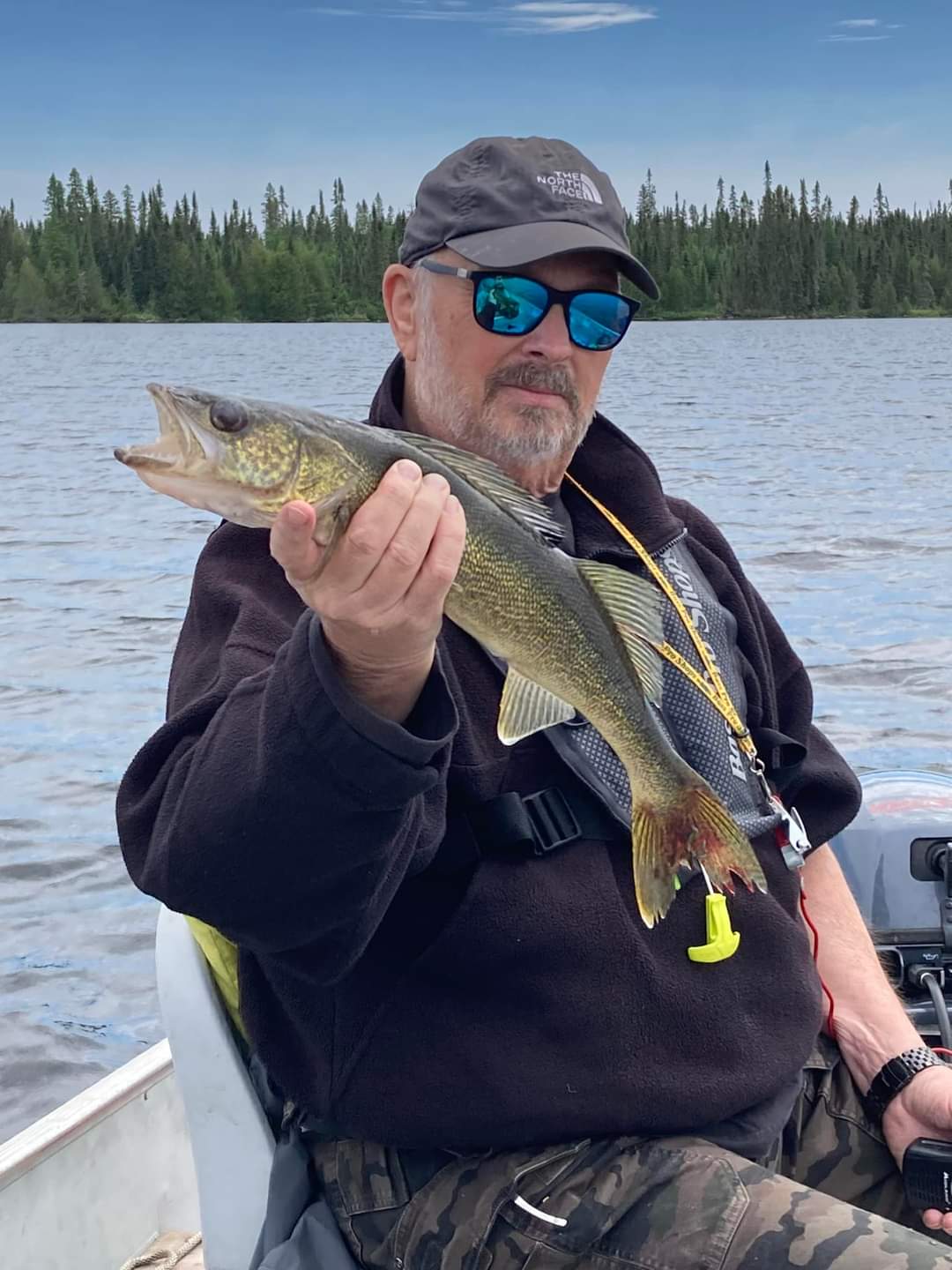 Angler in a cap and mirrored sunglasses holds up a walleye while seated in an aluminum boat on Pilot Lake.
