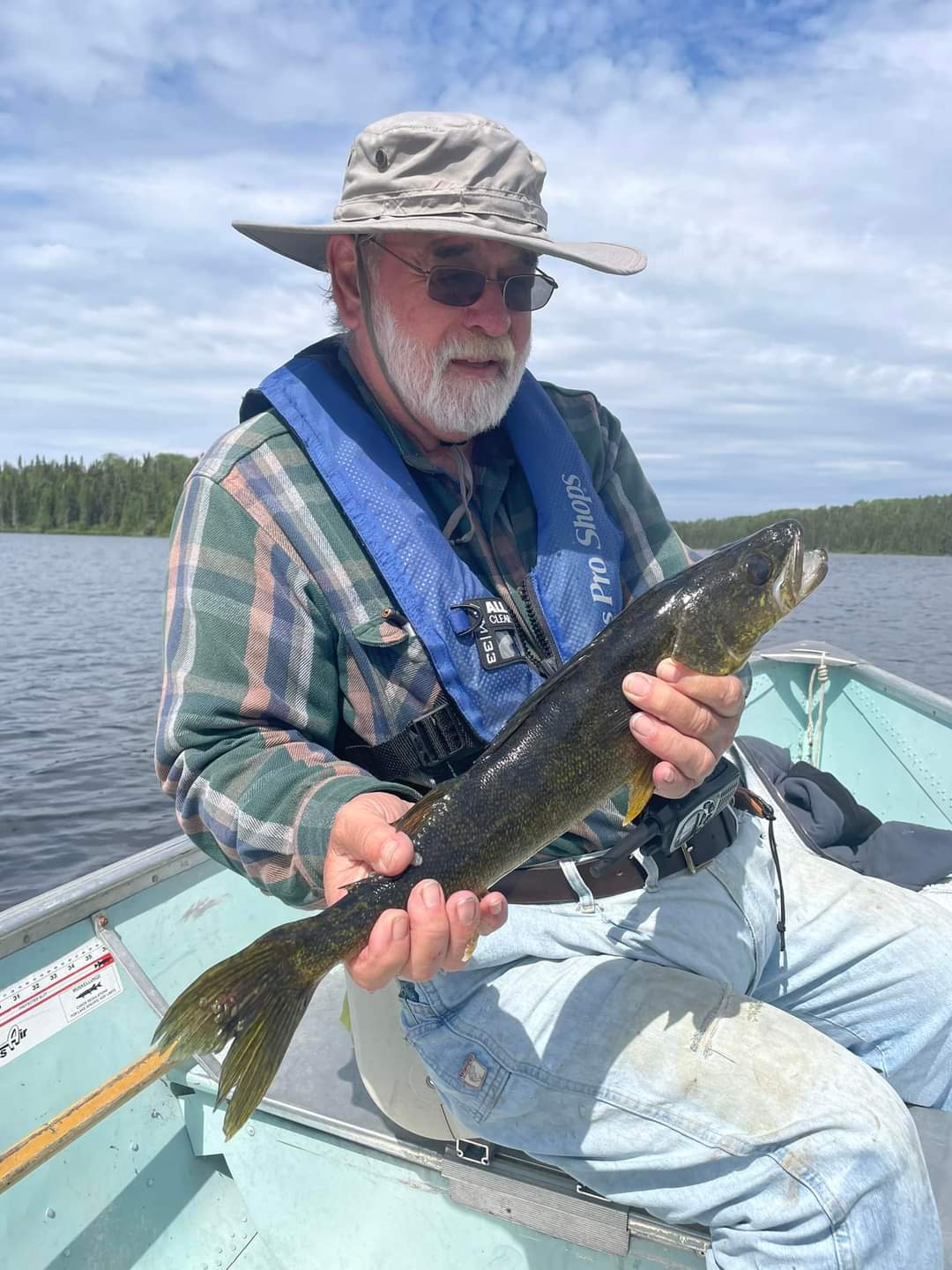 Angler in a wide-brim hat and life jacket holds a walleye in an aluminum boat on Pilot Lake, with forested shoreline behind.