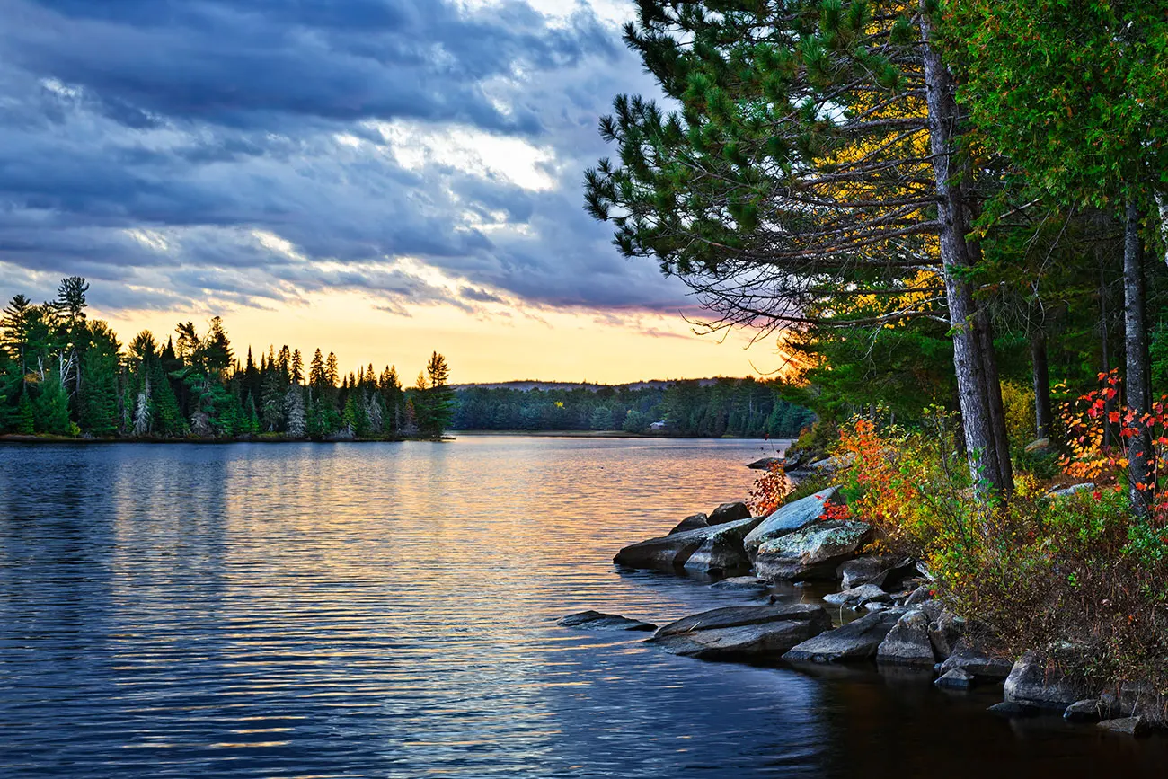 Pilot Lake at sunset, rocky shoreline with autumn foliage, tall pines, calm reflective water, and dramatic storm clouds lit gold.