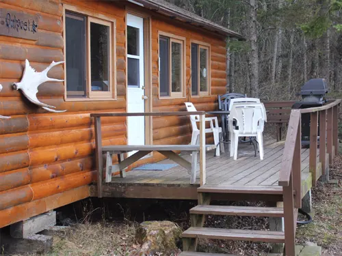 Log outpost cabin exterior with deck, white plastic chairs, moose antler wall mount, and surrounding boreal forest.