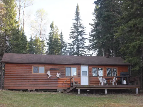 Rustic log outpost cabin with a front deck, patio chairs, and barbecue, surrounded by tall conifers on a grassy lot.