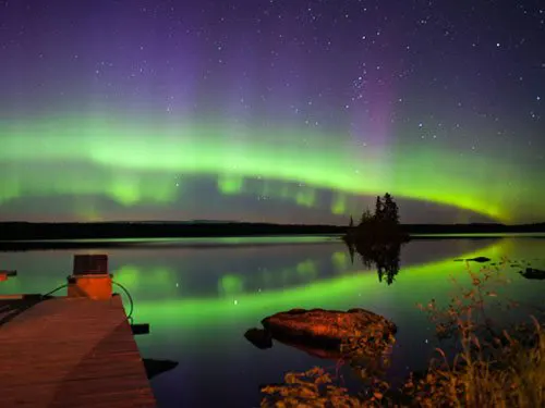 Aurora borealis in vivid green and purple arcs over Pilot Lake, reflected in still water beside a wooden dock and small island.