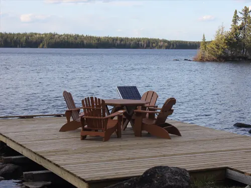 Adirondack chairs and a wooden table with a small solar panel on a dock overlooking Pilot Lake's calm waters and forested islands.