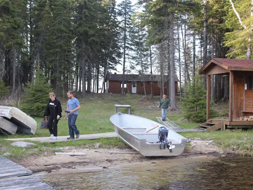 Three guests walk near the Pilot Lake shoreline beside two aluminum boats, with the outpost cabin and small outbuilding among the pines.