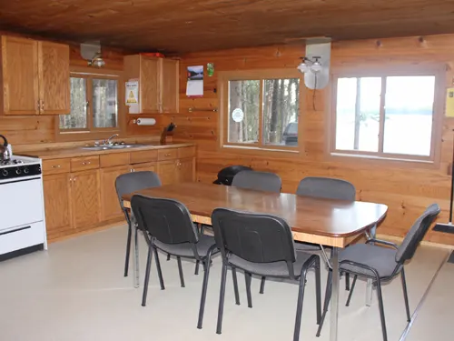 Bright dining area in the Pilot Lake outpost cabin, with a wooden table, six chairs, and a kitchen with oak cabinets and a white stove.