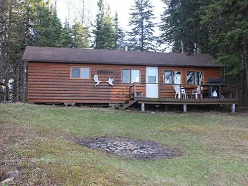 Log-sided outpost cabin with a large front deck, white patio chairs, and a barbecue, set among tall pines with a fire pit on the lawn.