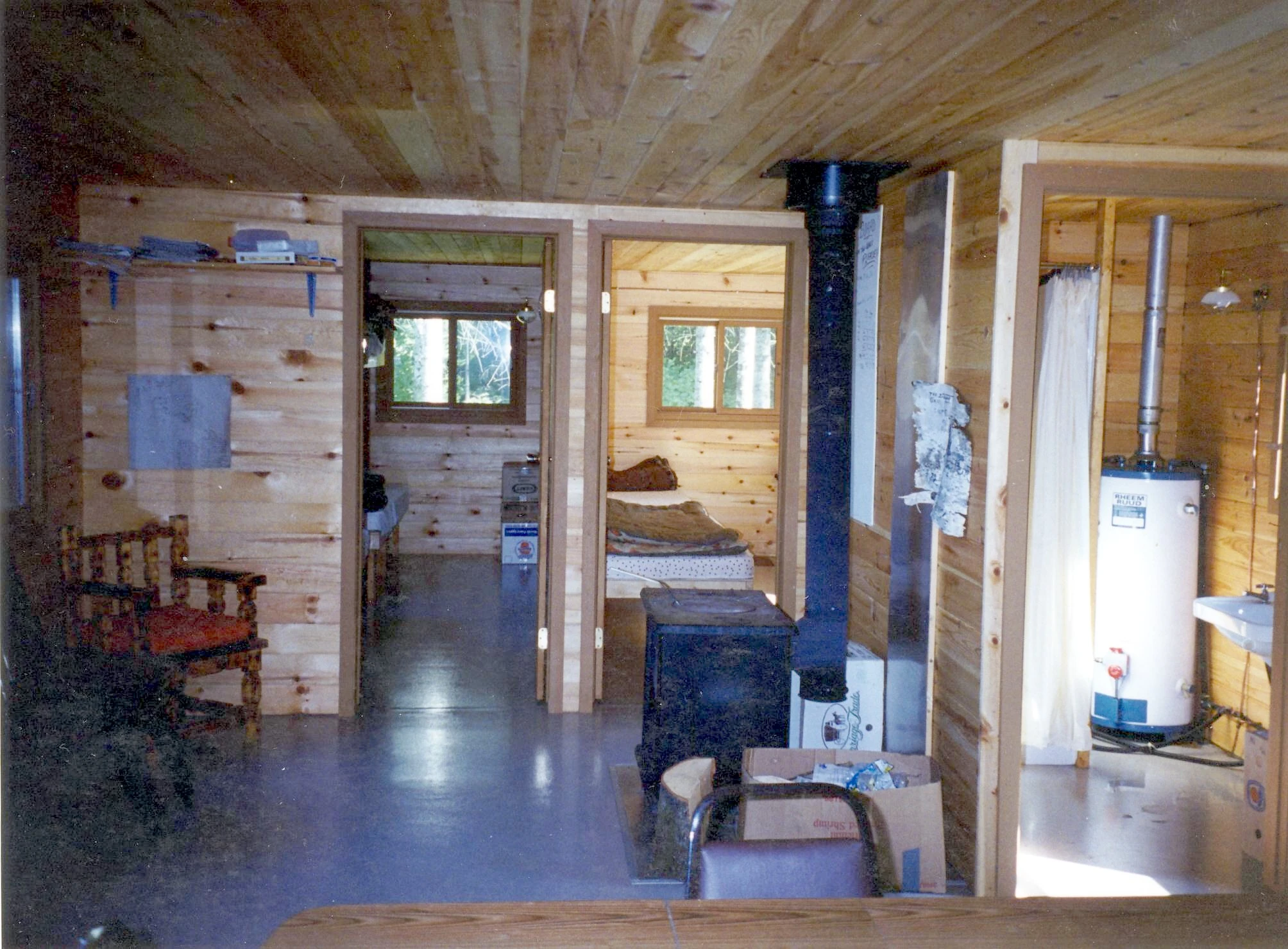 Interior of the Pilot Lake outpost cabin showing pine walls, a wood stove, water heater, two bedroom doorways, and a rustic chair.