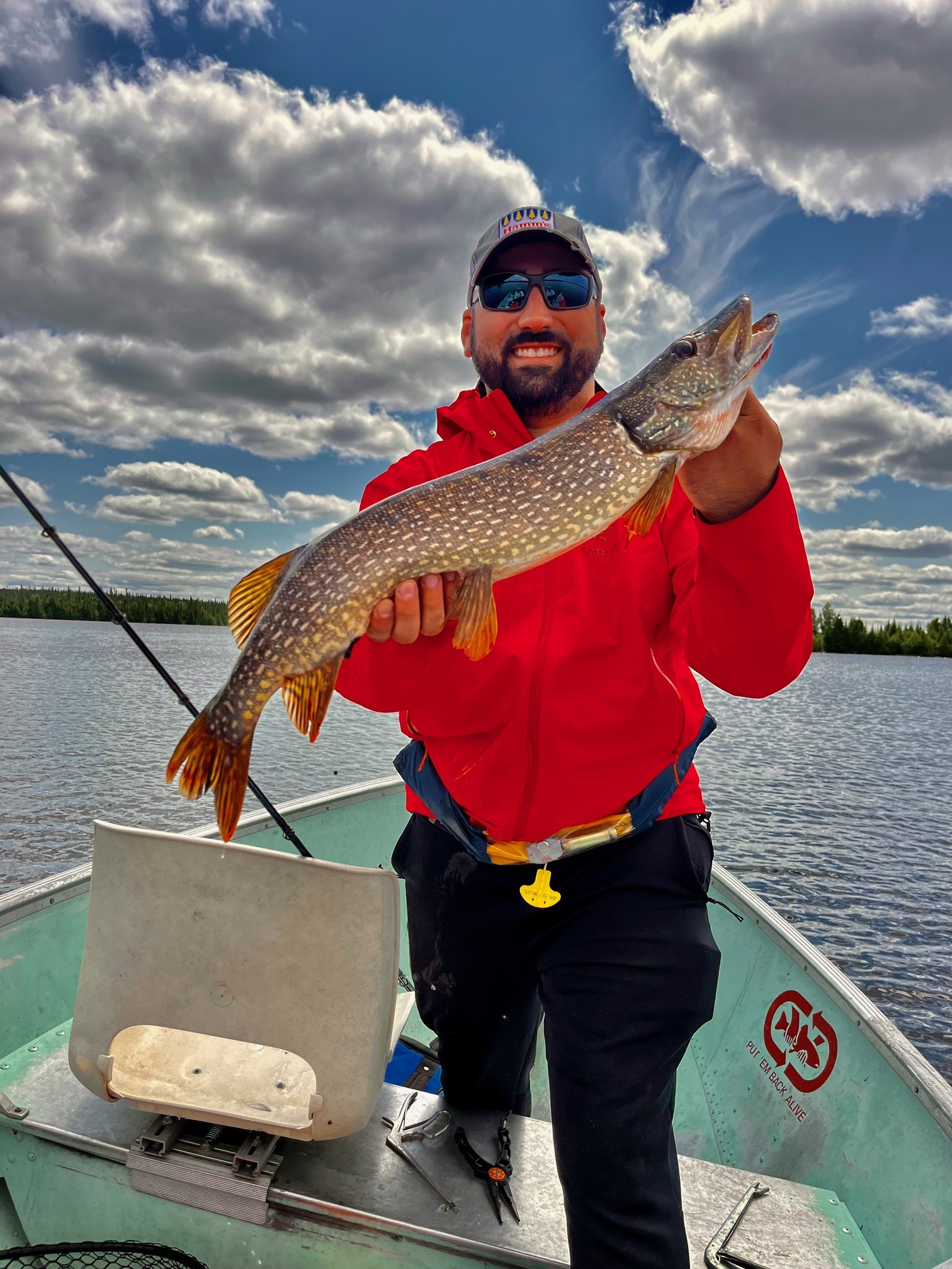 Smiling angler in a red jacket holds a northern pike aloft from a green aluminum boat on Pilot Lake under a cloudy sky.