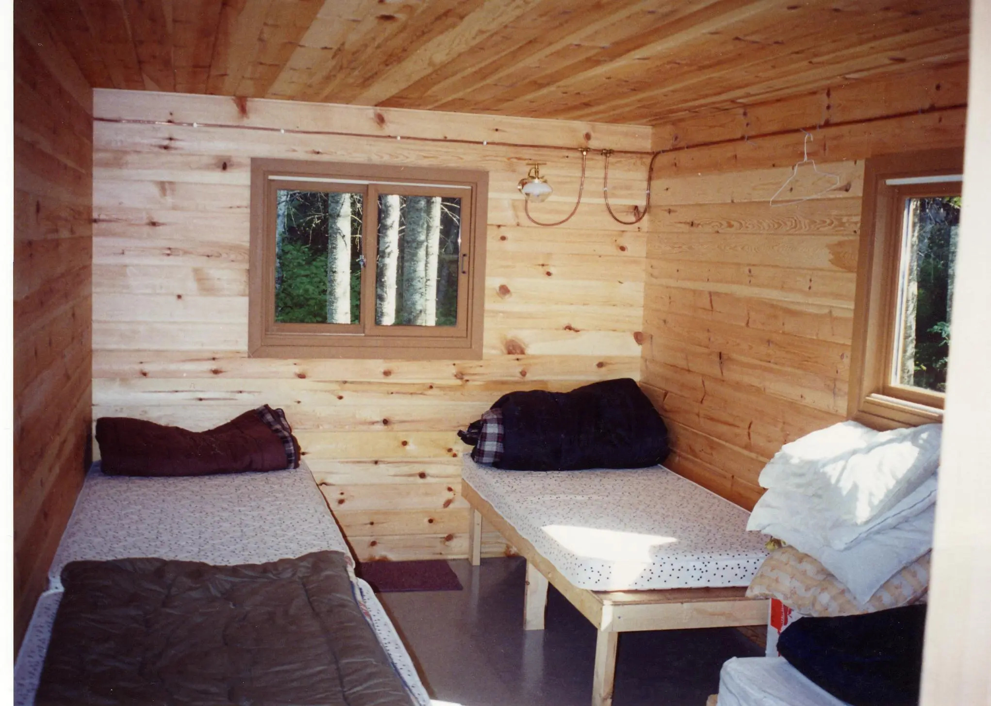 Interior of the Pilot Lake outpost cabin showing three single beds with sleeping bags and pillows, on knotty pine walls and ceiling.