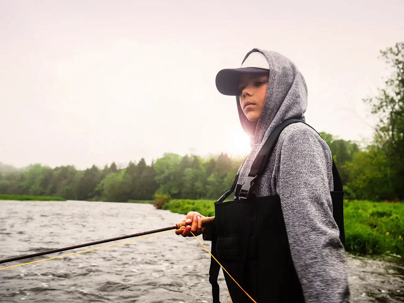 Young angler in hoodie and waders holds a fly rod streamside on a misty, overcast day at a remote Northern Ontario lake.