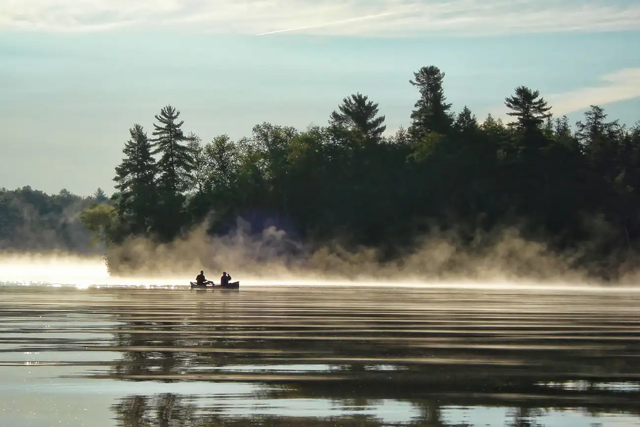 Two anglers in a canoe on a misty Sup Lake at dawn, pine-forested shoreline glowing behind rising morning mist.