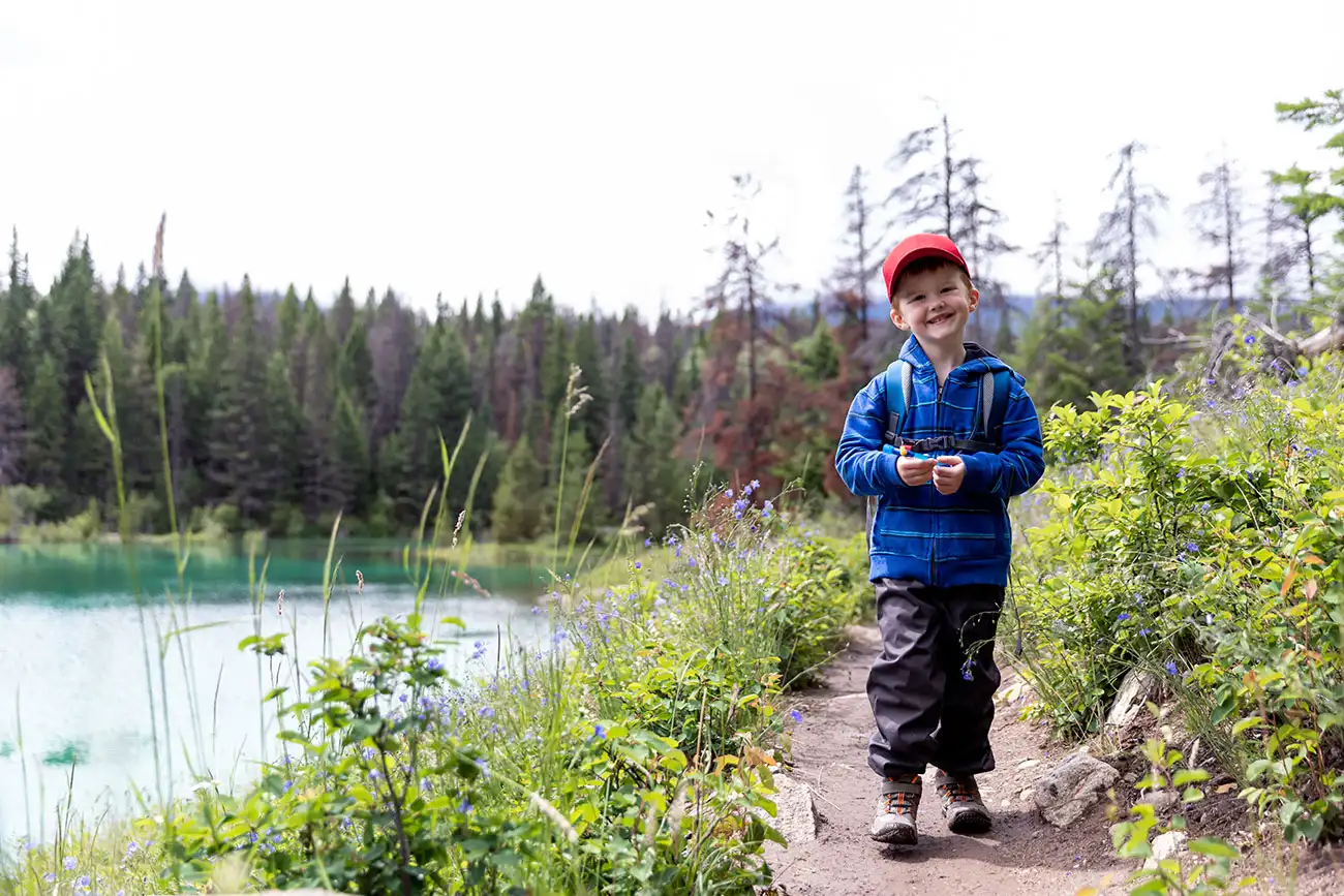 Young boy in red cap and blue jacket hiking a wildflower-lined trail beside a turquoise lake in Northwestern Ontario.