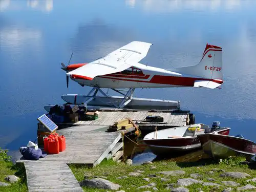 Red and white floatplane C-GFZF docked at a wooden dock on a calm Northern Ontario lake, with fuel cans and small boats nearby.