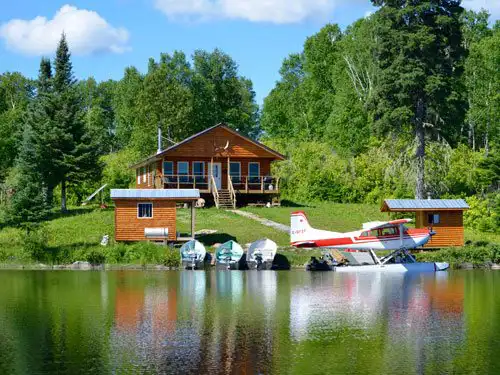 Red and white floatplane docked beside a log outpost cabin on a calm lake, with several aluminum boats beached nearby.