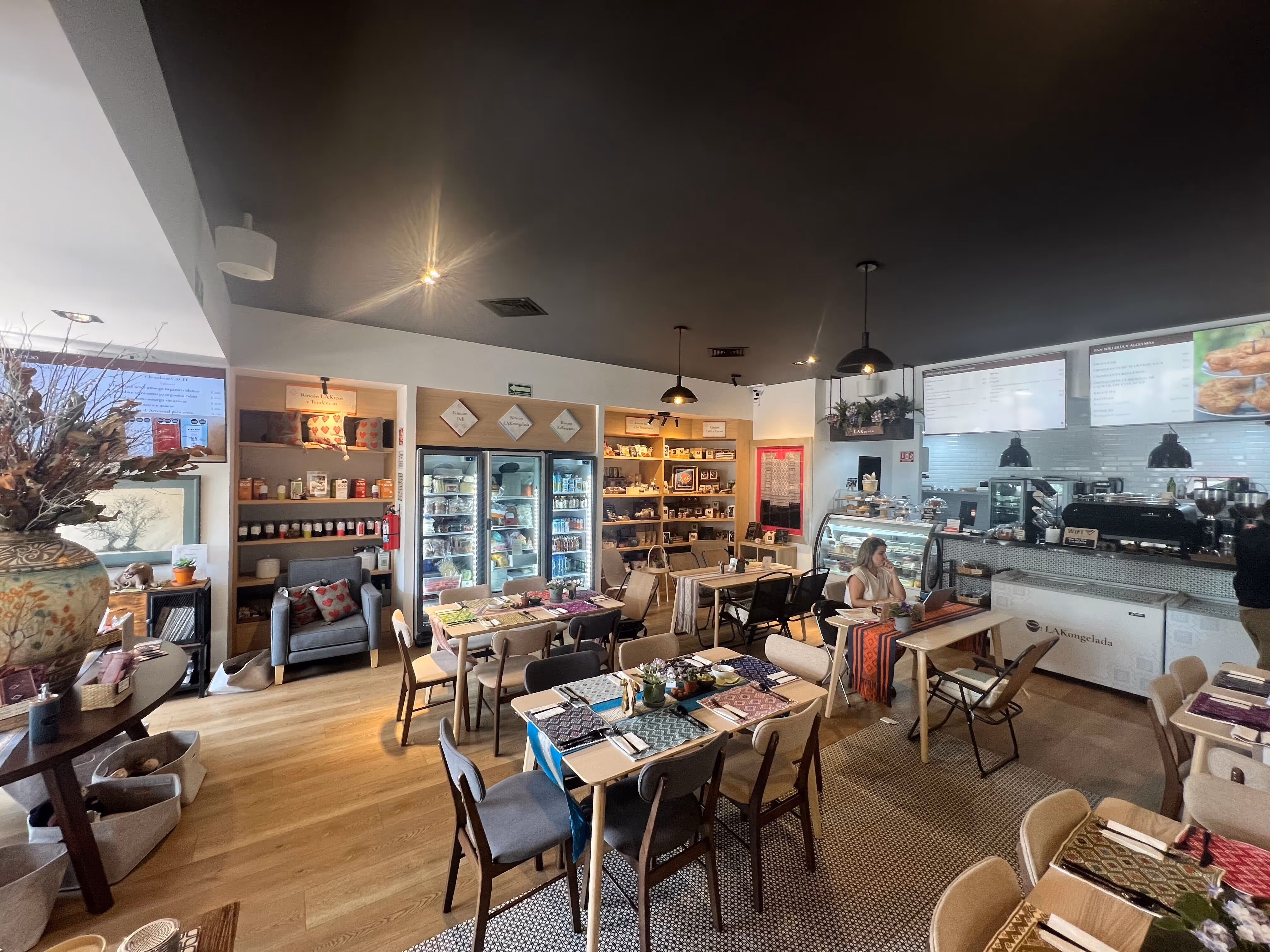 Interior de café luminoso con mesas, sillas, refrigeradores con productos y una persona sentada cerca del mostrador.