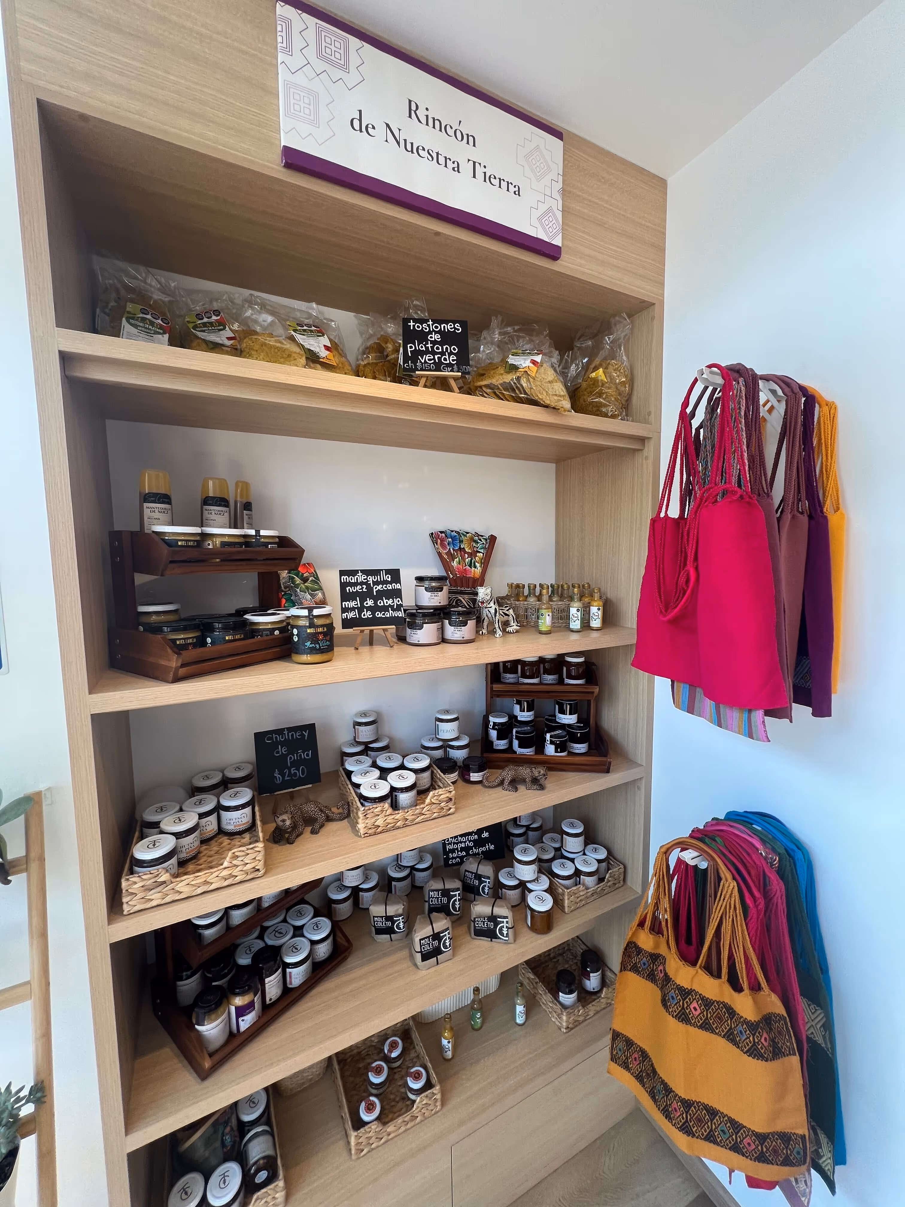 Estante de madera con productos artesanales y bolsas coloridas colgadas a la derecha, bajo un letrero que dice 'Rincón de Nuestra Tierra'.