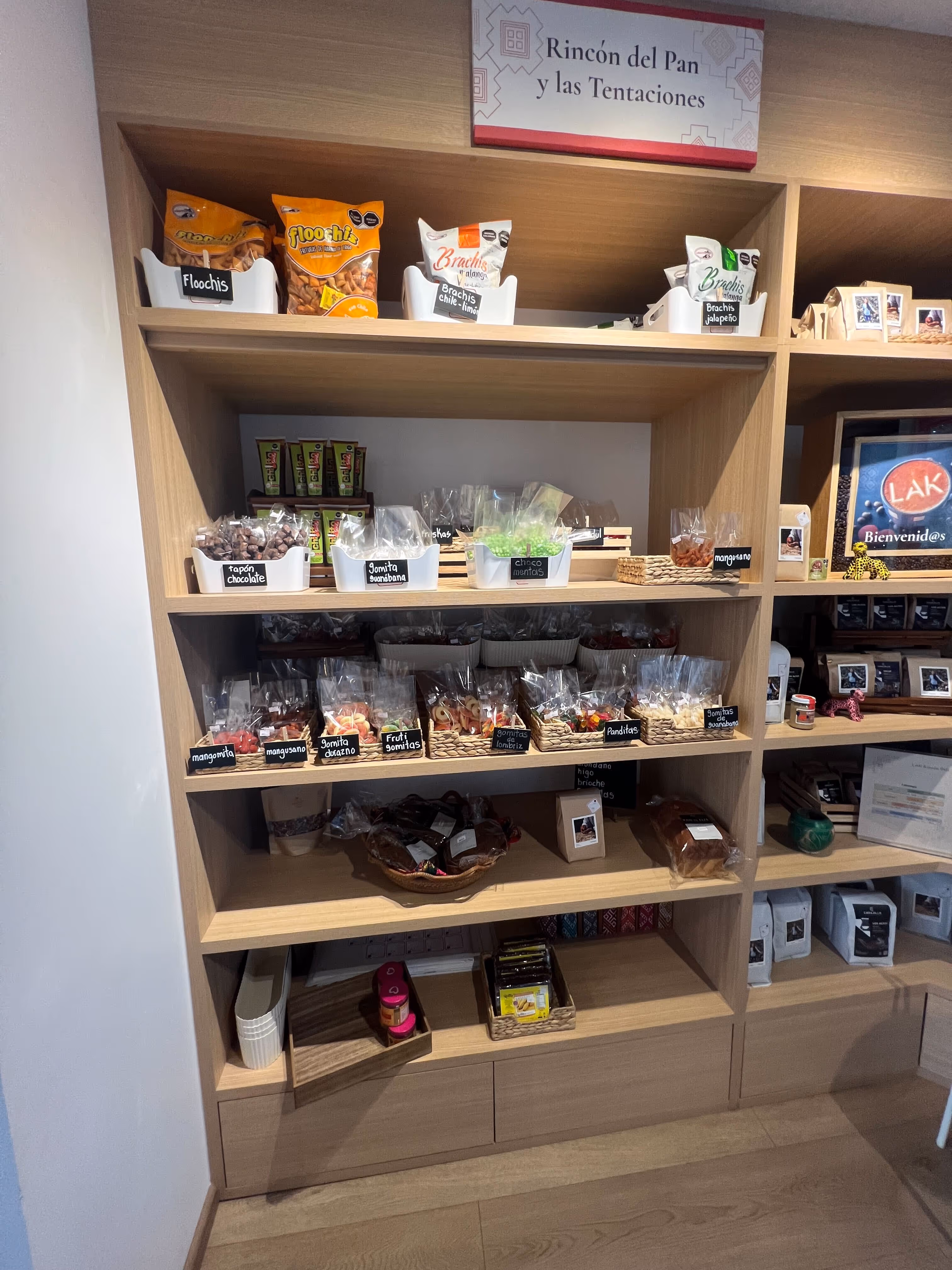 Estantería de madera con diferentes botanas, dulces y productos etiquetados bajo un letrero que dice Rincón del Pan y las Tentaciones.