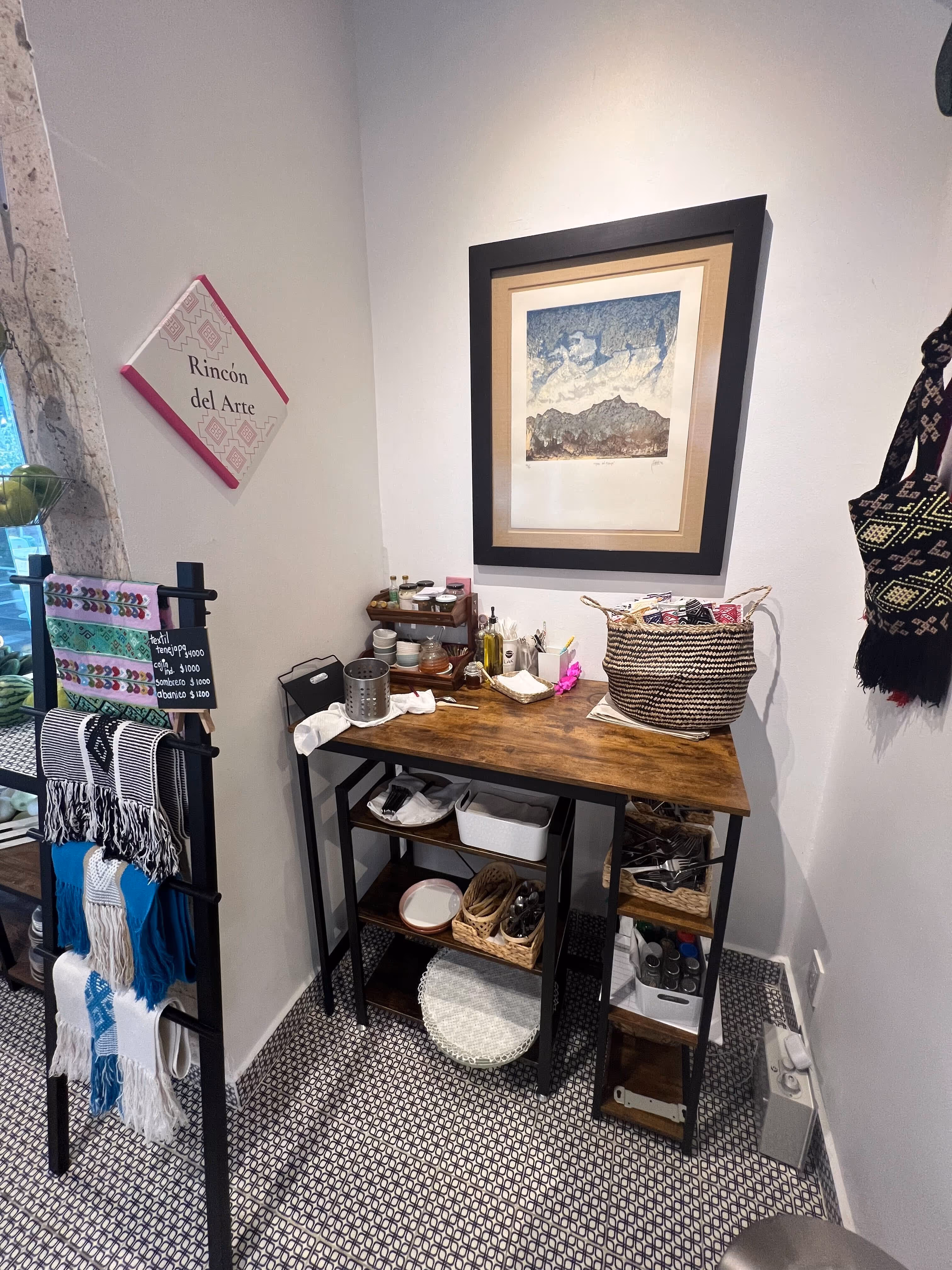 Esquina de una tienda de arte con mesa de madera que sostiene una canasta y utensilios, paredes blancas, un cuadro enmarcado y una exhibición de textiles coloridos.