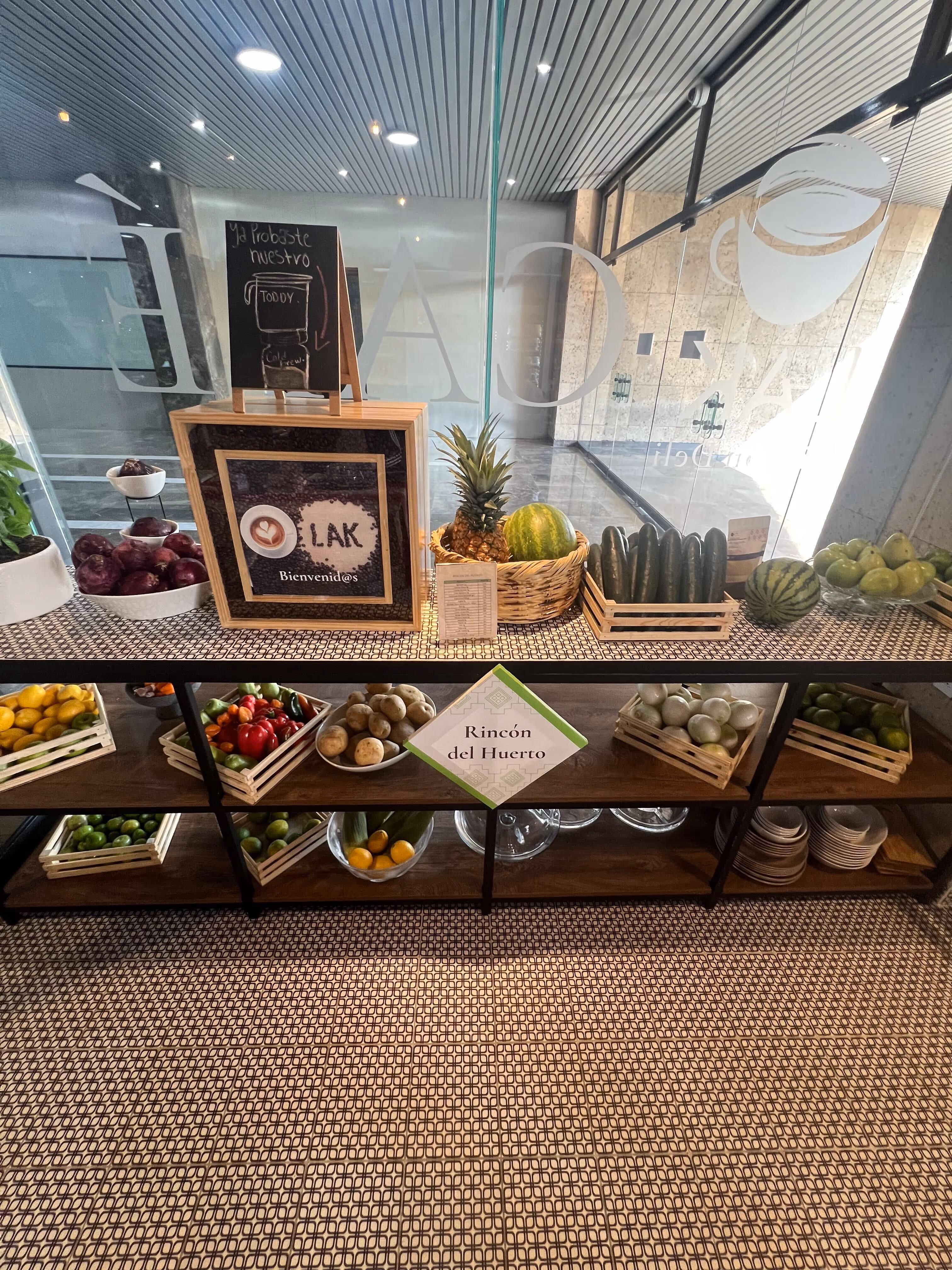 Estante de madera con frutas y verduras frescas organizadas en cestas, con un letrero que dice ‘Rincón del Huerto’ en un espacio moderno con paredes de vidrio.