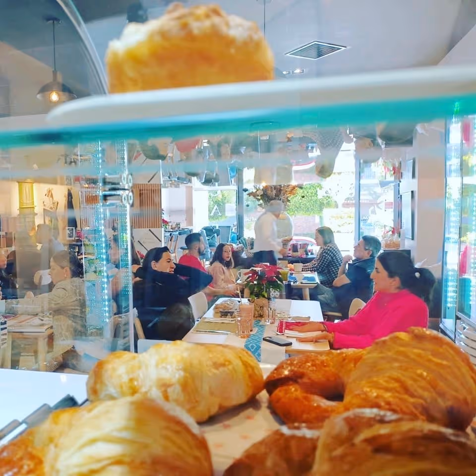 Variedad de panes y bocadillos en primer plano dentro de un exhibidor de vidrio, con personas conversando y comiendo en un café al fondo.