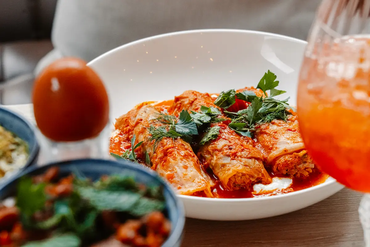 Close-up of herbal cabbage rolls in sauce next to a vibrant orange spritz cocktail.