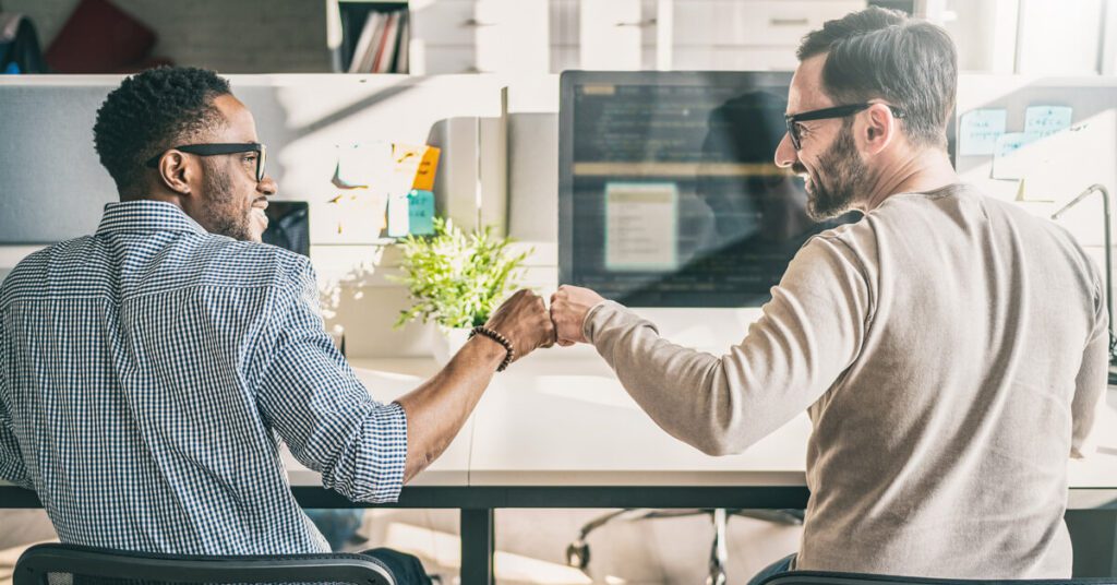 Two coworkers fist-bumping at a shared desk with computers.