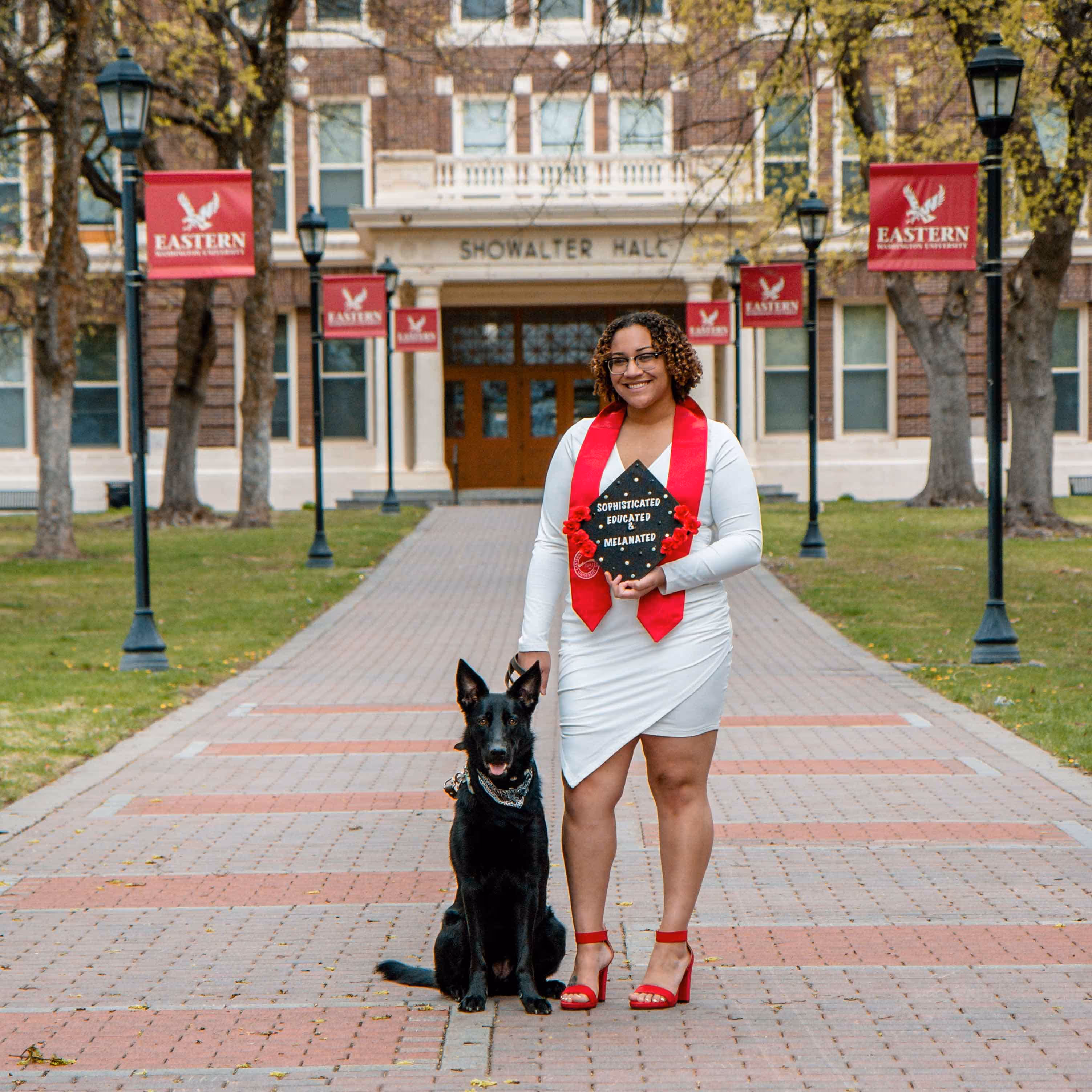 Smiling graduate standing next to a black dog in front of Showalter Hall at Eastern Washington University.