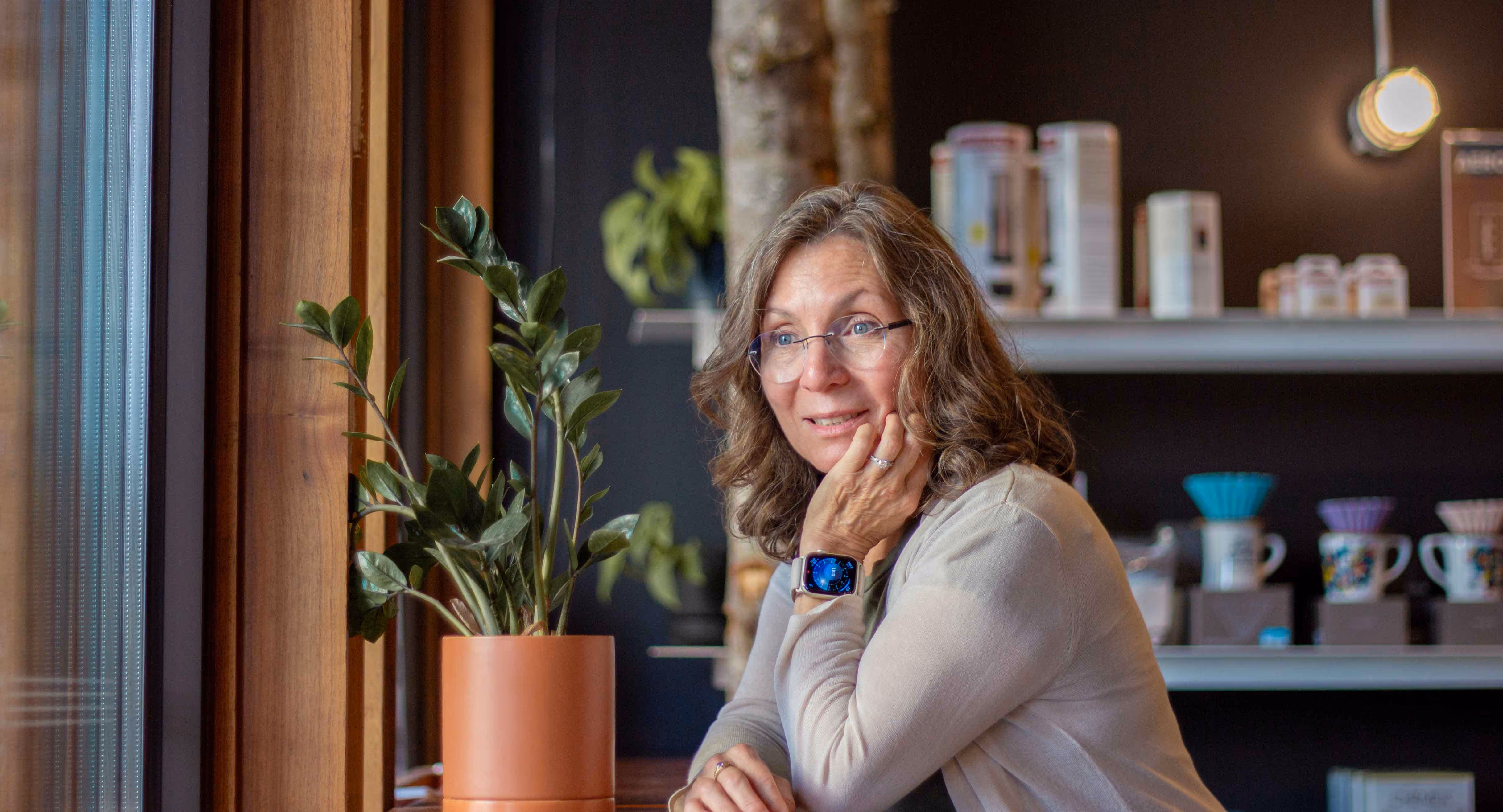 Middle-aged woman sitting indoors by a wooden window, resting her chin on her hand and smiling.
