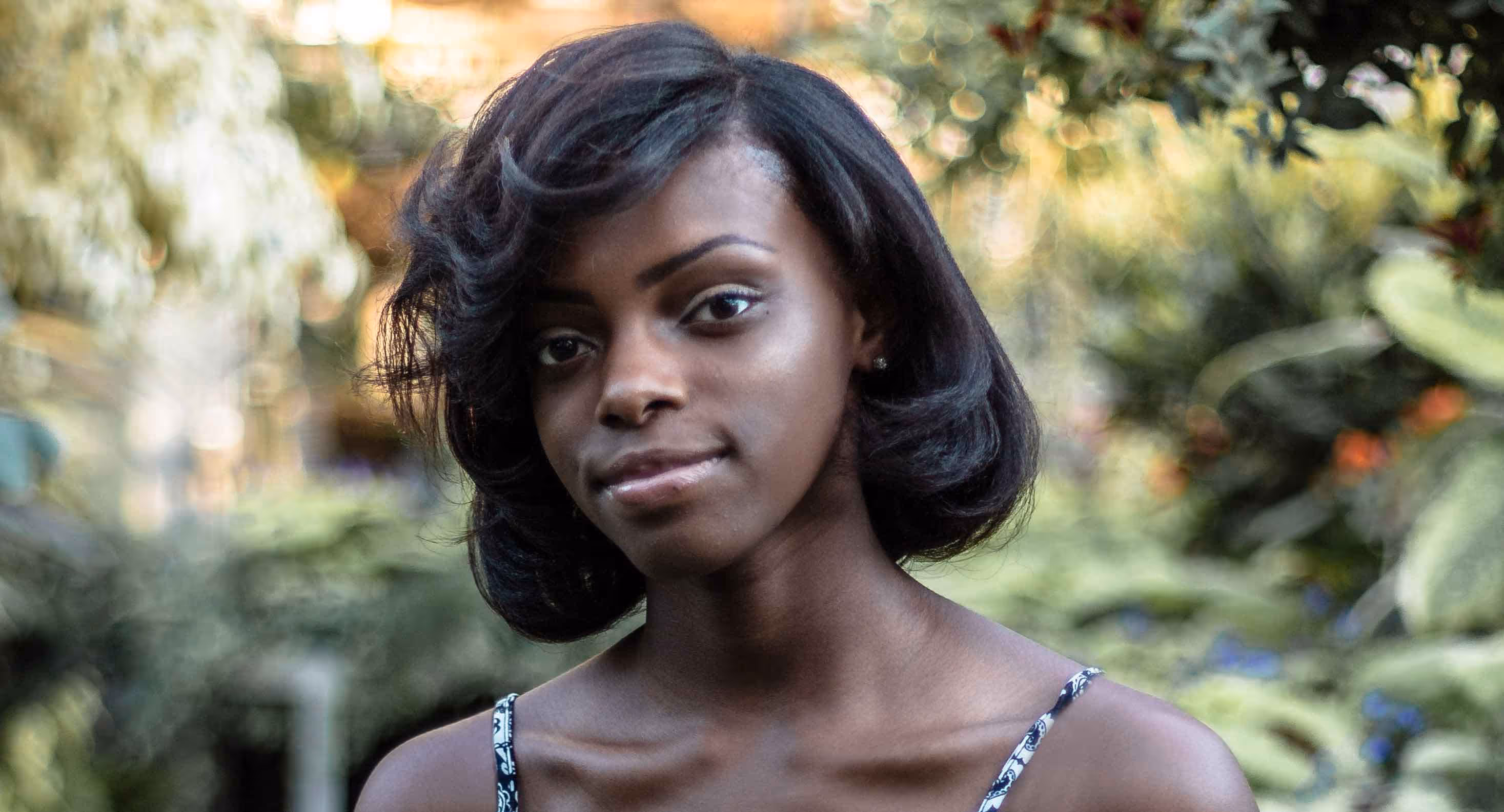 Portrait of a young woman with short black hair standing outdoors with blurred greenery in the background.