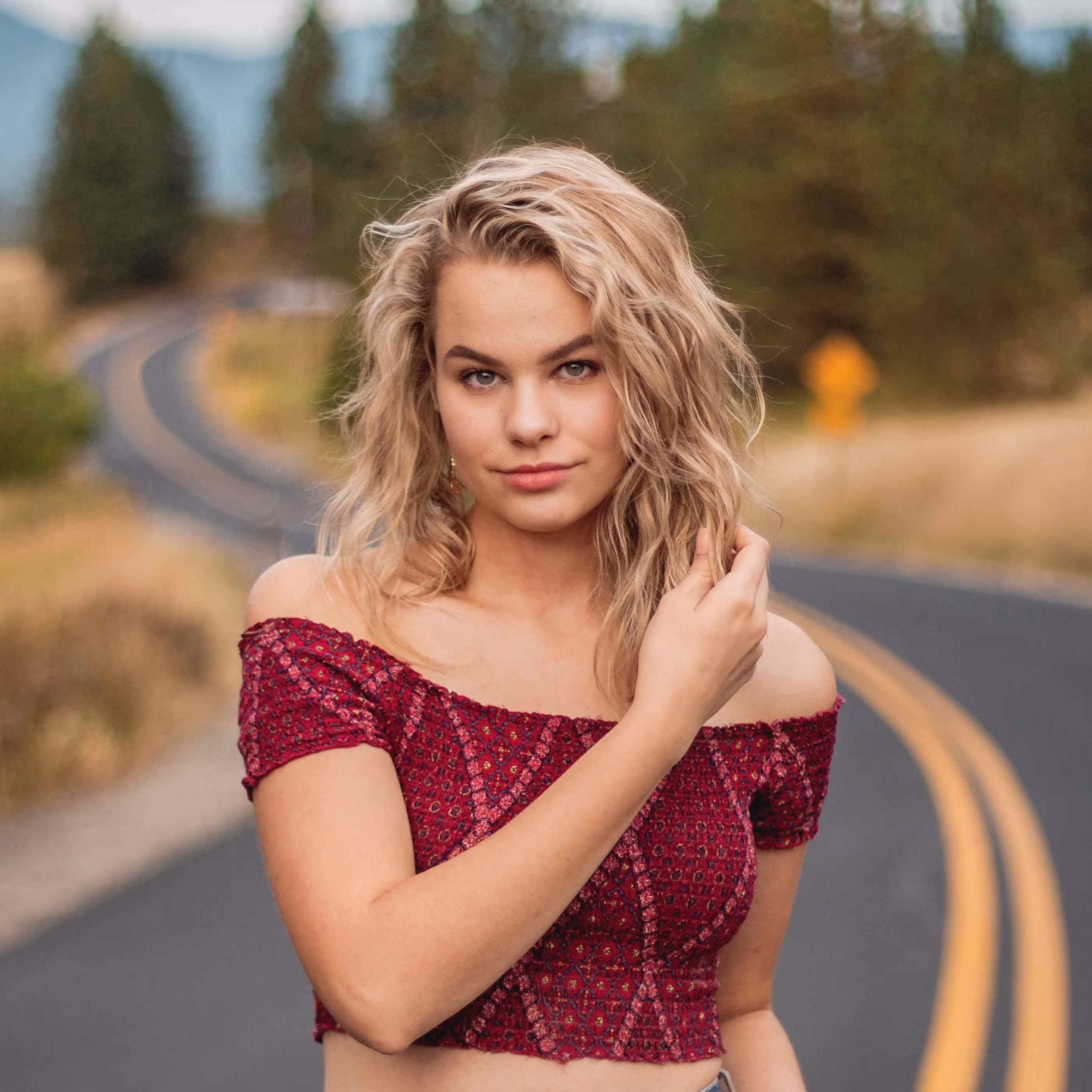 Blonde woman with wavy hair wearing a red off-shoulder top stands on a winding road with trees in the background.