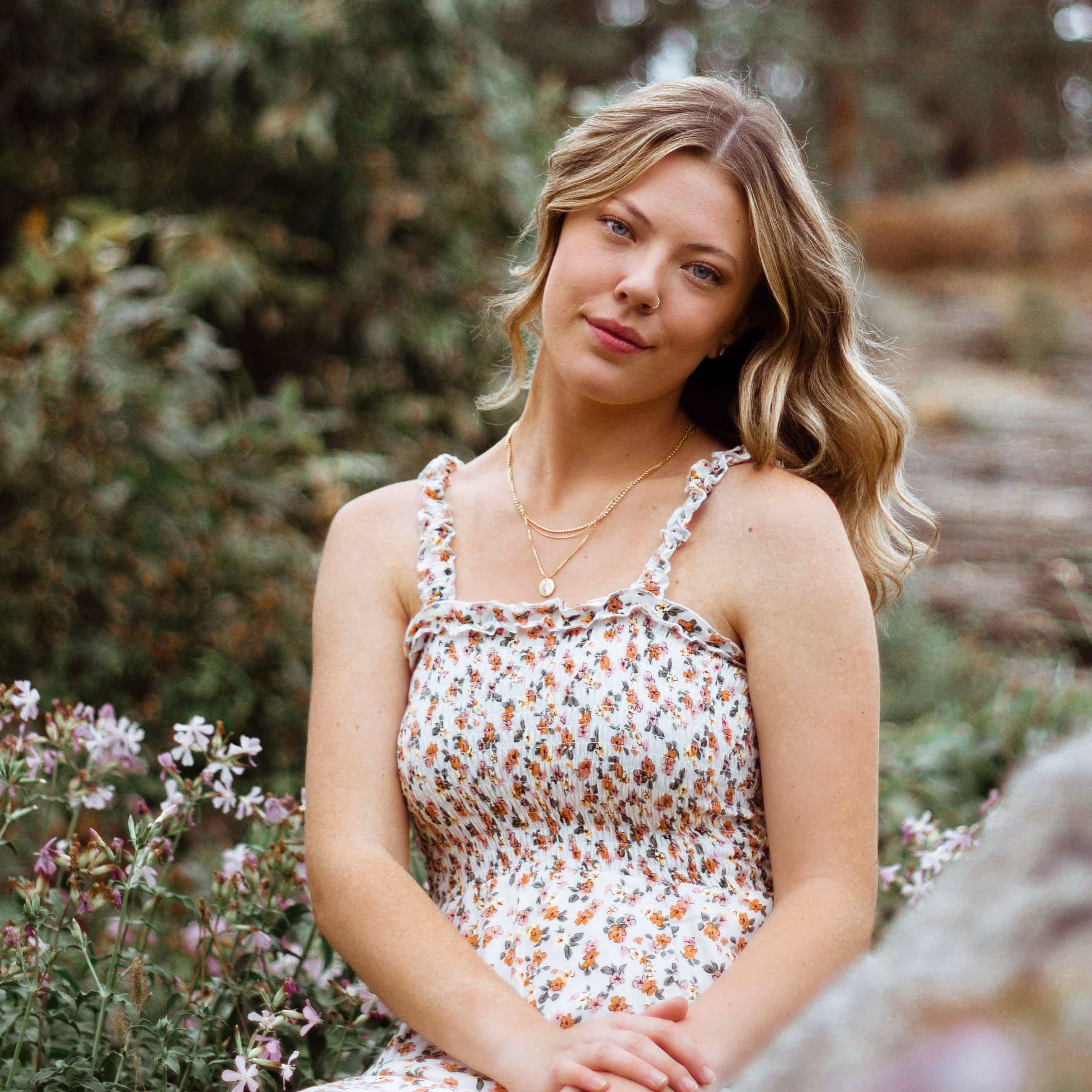 Young woman with blonde wavy hair wearing a white floral dress sitting outdoors amidst greenery and flowers.