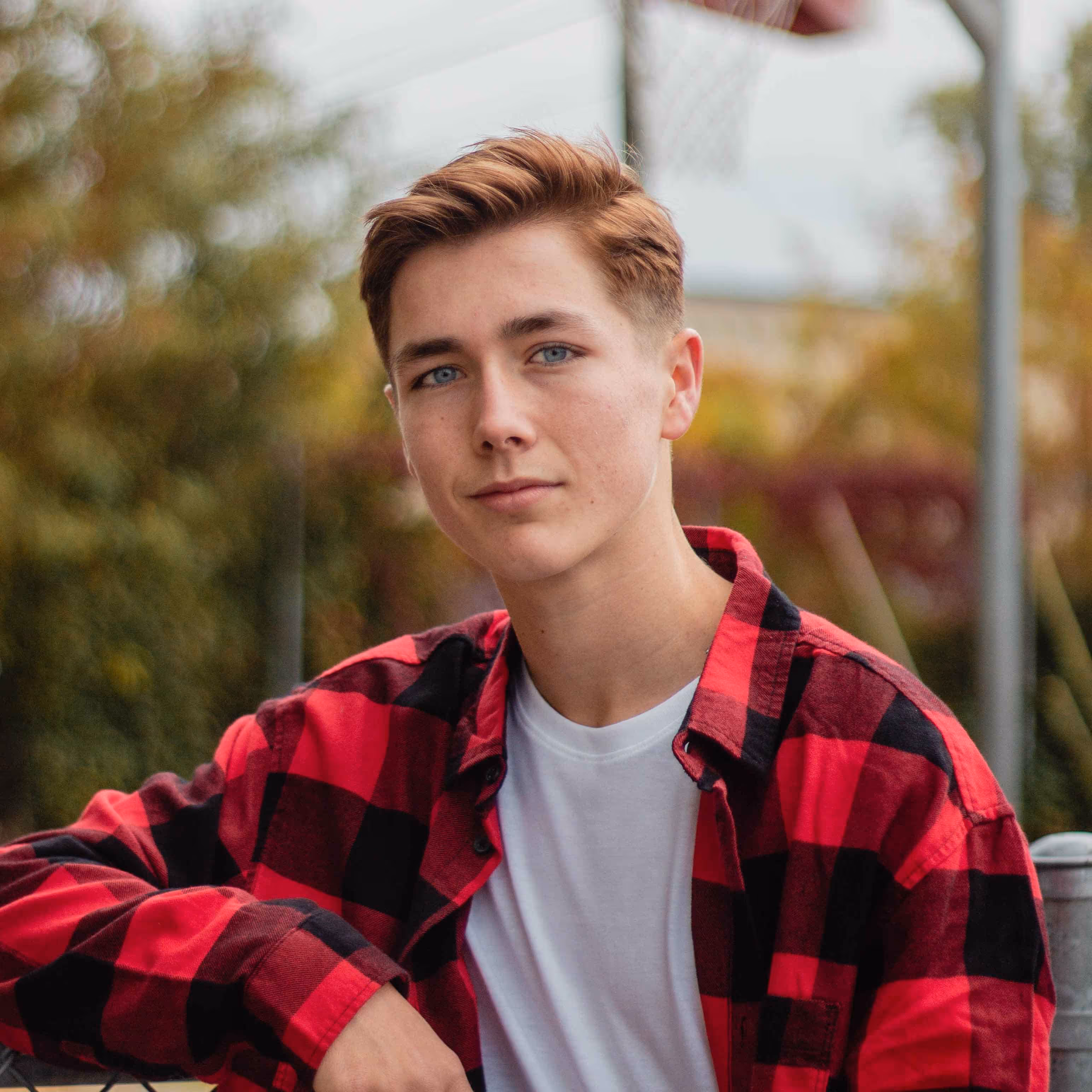 Young man in a red and black checkered shirt posing outdoors near a basketball hoop.